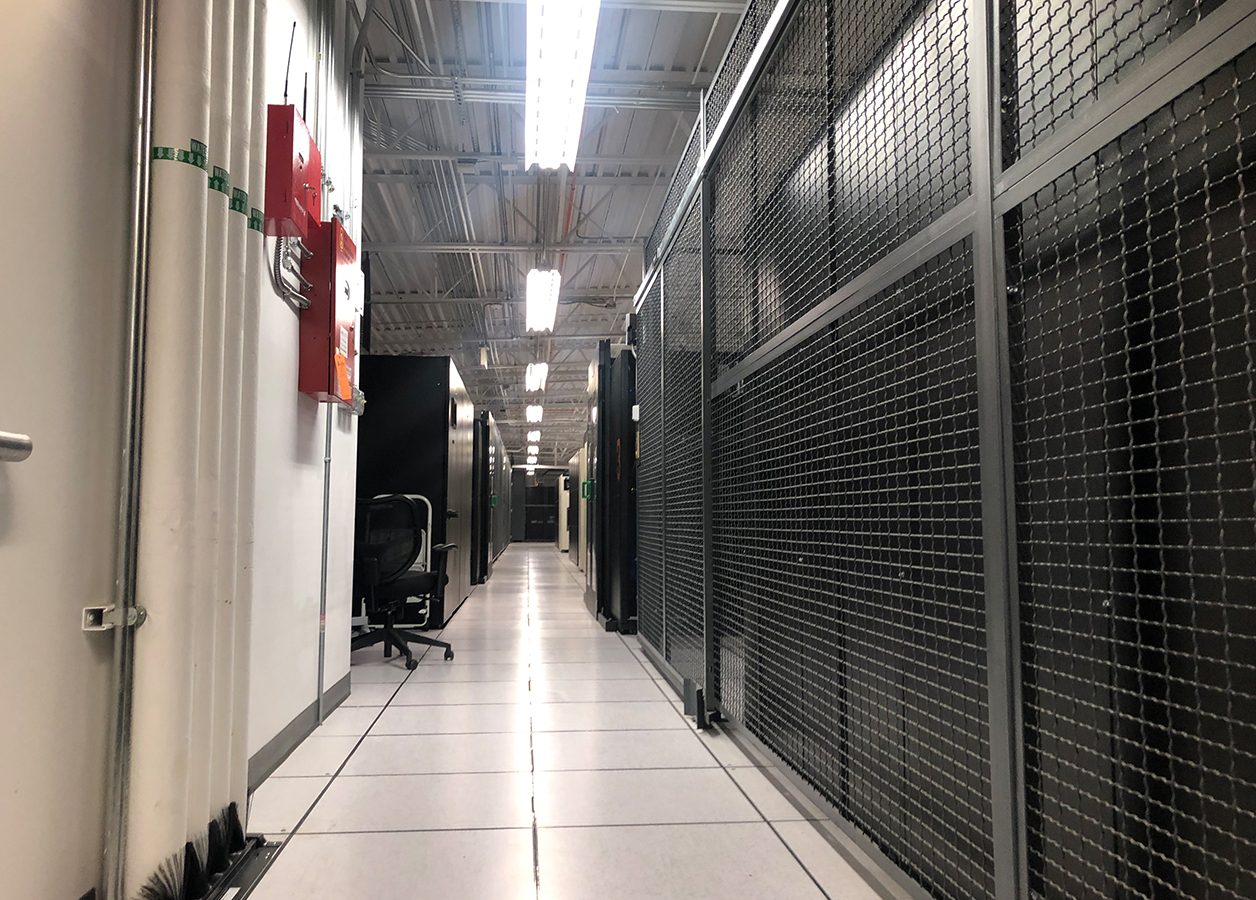 The inside of a datacenter. On the left, pipes, presumably for coolant, can be seen. On the right, wire mesh cages enclose datacenter racks (though they are hard to see). A lit aisle between datacenter equipment stretches into the distance.