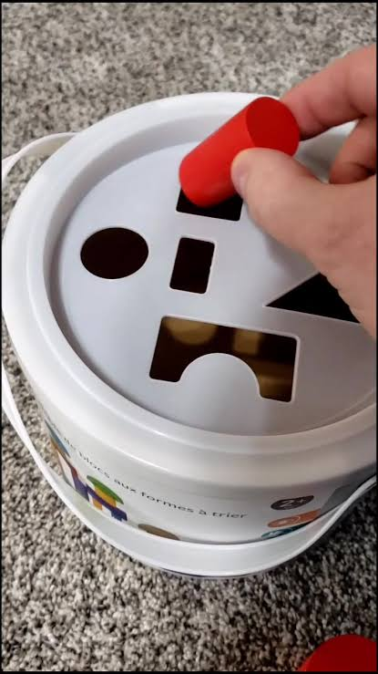 Picture of a sorting toy. The hand of the person framed in the picture holds a red cylinder just above the square hole, which just so happens to fit perfectly.