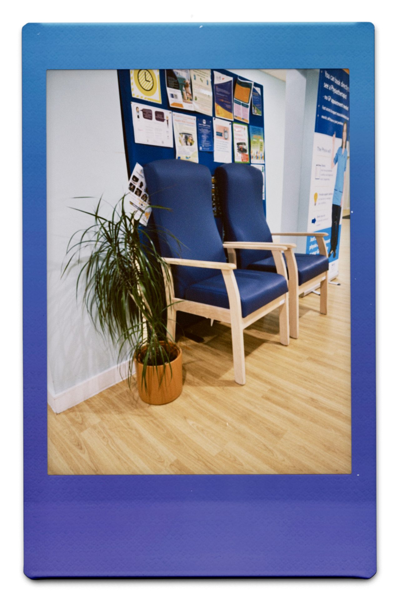 Image shows the GP waiting room, with two, blue, high-backed chairs behind which sits a board of various medical posters.
The floor is a light coloured, laminated wood. To the right of the chairs (left in the image) is a small potted plant.