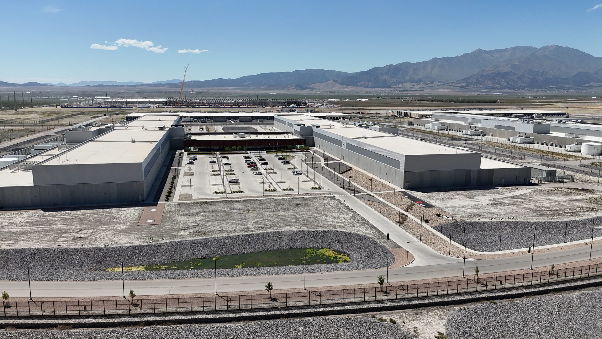 A massive campus stretches out into the distance. In the foreground, two large, long buildings can be seen, surrounded by generation and cooling equipment. The whole campus is ringed by a road.