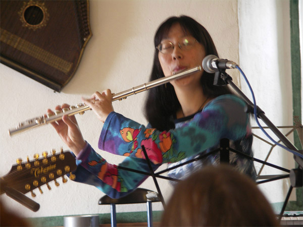 Debbie playing the flute at a microphone. She is wearing a colourful top. You can see part of a guitar to the left.